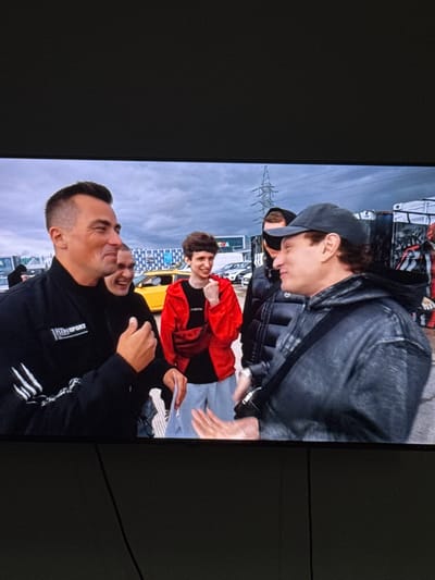A group of five men are gathered outdoors, seemingly at an event. The man in the foreground on the right wears a dark grey hooded jacket and a grey baseball cap. To his left, a man in a black jacket with white stripes on the sleeves smiles broadly. Behind him, another man laughs. To the right of the laughing man, a man in a red jacket stands with his hands in his pockets, holding a small piece of paper. In the background, a yellow car is visible, along with other vehicles and structures, including a large power pylon and a building with graffiti. The sky is overcast with grey clouds. This scene does not appear to be located in Neubrandenburg, Germany, as there are no identifiable landmarks or contextual clues to suggest this specific location.