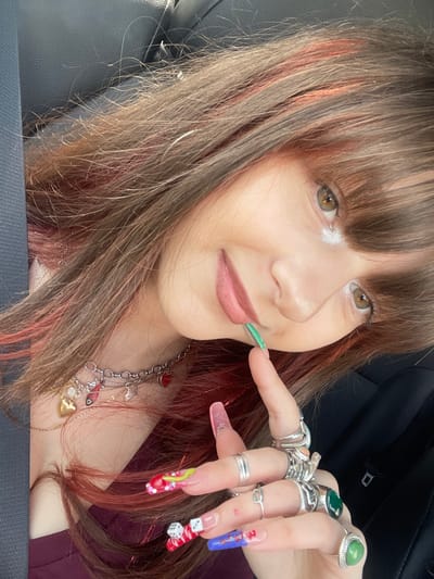 The image depicts a female individual in a close-up, angled portrait taken inside a vehicle in Kfar Saba, Israel. The subject, viewed from the upper torso, has brown hair with reddish-orange highlights and bangs, and is looking directly towards the camera. Her eyes are hazel-brown with white glitter applied to the inner corners. Her lips are covered with a pink-toned lip product. She is wearing a dark red or maroon garment. Multiple silver-toned rings adorn the fingers of her visible right hand, including rings with dark green and light green stones. Her nails are long, artificial, and elaborately decorated: the thumb nail is green, the index nail is light pink/clear, the middle nail is red with yellow lines, green accents, and rhinestones, the ring nail is white with red splatters and small white dice, and the pinky nail is blue with red splatters and white markings. A silver-toned chain necklace with multiple charms, including gold and red hearts, is visible around her neck, along with a thinner gold-toned chain. Her right index finger is positioned near the corner of her mouth. The background consists of dark-colored, textured upholstery, consistent with a car seat interior.