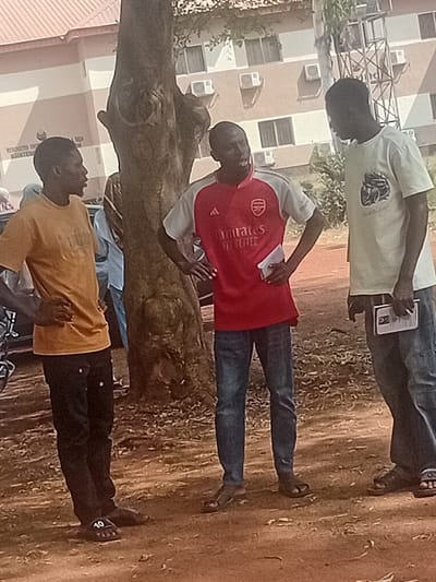 Three men stand outdoors near a building with many air conditioning units. The ground appears to be reddish-brown soil. One man wears a red Arsenal football jersey, while another has on a light tan shirt and jeans while holding a paper. The third man is wearing a yellow-gold shirt with black pants and sandals. They are near a large, bare tree trunk.