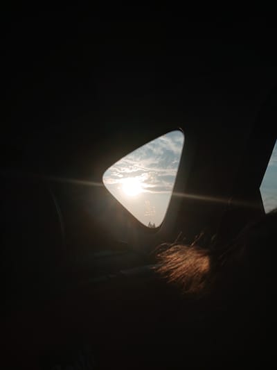 An interior view from the passenger side of a vehicle looking out a small, triangular side window. The sun is setting, casting a strong glare and lens flare across the sky, which is filled with scattered clouds. The interior of the vehicle is dark, with a portion of a person's head and hair visible in the foreground, illuminated by the setting sun. There is text visible on the window, including what appears to be a car manufacturer's logo. The overall impression is of a travel scene during sunset, looking out from the back of the car. The location of Novokudashevo, Russia, is mentioned as a potential contextual detail, but cannot be visually verified from the image itself.