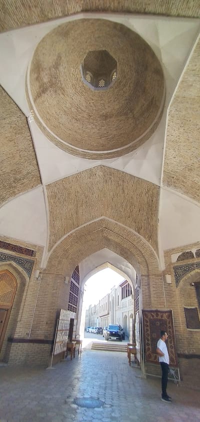 This is a low-angle, full-shot, vertical, outdoor photograph taken in Bukhara, Uzbekistan. The image features a domed ceiling constructed of bricks, with geometric patterns. The dome opens into an octagonal structure with small, window-like openings. Below the dome, arched passageways lead to a street scene. In the foreground, a man in a white t-shirt and black pants stands facing left, looking into the distance. He is positioned near several large rugs displayed vertically. The passageway opens to a street with a line of parked cars and buildings on either side. The ground is paved with cobblestones. The architectural style suggests a historical site, likely a covered bazaar or a madrasa within Bukhara's old city.