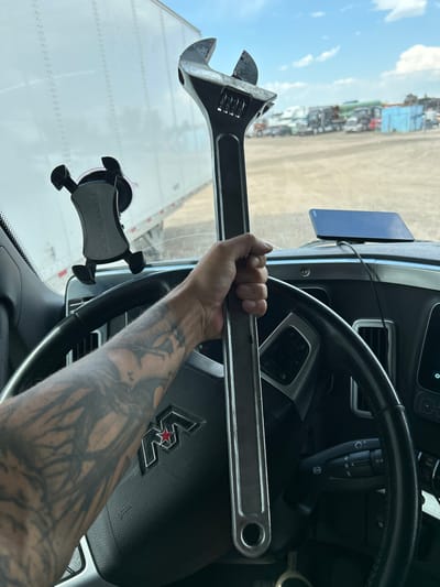 The image shows a close-up view from inside a truck cab, looking out the windshield. A large adjustable wrench is being held by a person's left arm, which is heavily tattooed. The wrench is positioned vertically in the frame, obscuring part of the view outside.

Outside the truck, parked in what appears to be a lot or yard, are several other trucks and trailers, suggesting a commercial vehicle depot or trucking hub. The sky is visible with some clouds. A phone mount is attached to the windshield, and a phone is visible on the dashboard. The steering wheel of the truck is in the foreground.

The scene is likely located in Aurora, United States of America, given the context of commercial trucking operations. The environment appears to be outdoors during the daytime, with bright lighting. The overall impression is one of a working environment, possibly related to truck maintenance or operations.