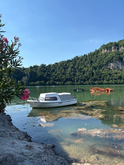 The image depicts a serene lake scene with a clear blue sky and lush green hills in the background. In the foreground, a white motorboat is moored close to a rocky shore, its reflection shimmering on the calm water. To the right, an orange paddle boat is visible on the lake. The water is a clear greenish hue, with some submerged vegetation and debris visible near the shore. The presence of these boats and the natural landscape suggests a recreational area, possibly a tourist destination. The image captures a peaceful day at the lake, with no people actively engaged in activities.