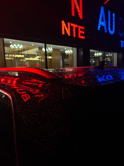 A nighttime shot features a car's exterior, wet with raindrops that reflect red and blue neon signage, "NOTE AU" vertically. A building is reflected in the car's surface, showing brightly lit interiors with chandeliers and horizontal wooden beams. The car is mostly in darkness, with only portions illuminated by the colorful light. The overall scene is abstract, with the reflections creating a distorted and fragmented view.