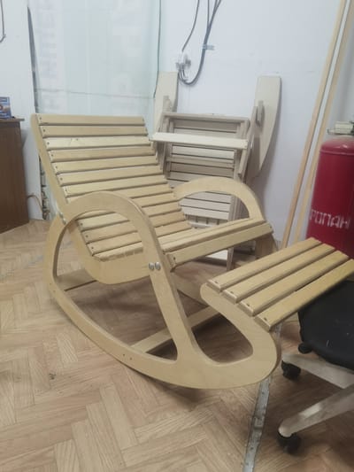 A wooden rocking chair with a detached footrest sits on a parquet floor. The chair is made of light-colored wood with a slatted seat and back. The footrest is also slatted and positioned in front of the chair. In the background, a foldable wooden chair is leaning against a wall. To the right, a tall red cylinder with white Cyrillic text that appears to read "ПОЖАРНЫЙ" (firefighting) is visible. The setting appears to be an indoor workshop or storage area, with exposed electrical wiring and a metal strip on the floor. There are no people present, and the overall mood is still and unposed. The lighting suggests an indoor environment, possibly during the daytime. The location is specified as Cherkessk, Russia.