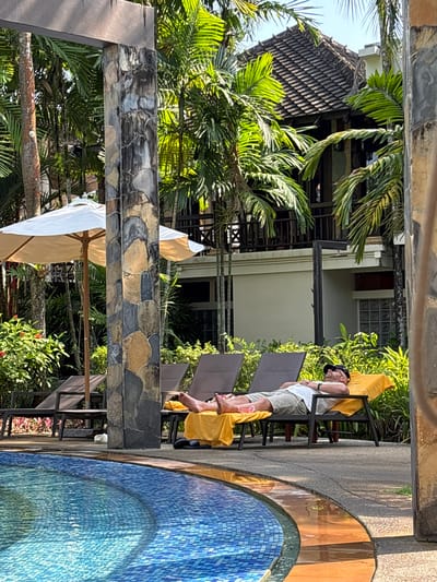 A man is sleeping on a lounge chair next to a swimming pool in Khok Kloi, Thailand. He is wearing a white shirt, khaki shorts, and a black baseball cap. The pool is tiled with blue and yellow mosaic tiles. Palm trees and a building with a brown roof are in the background. An umbrella is partially visible in the lower left corner.