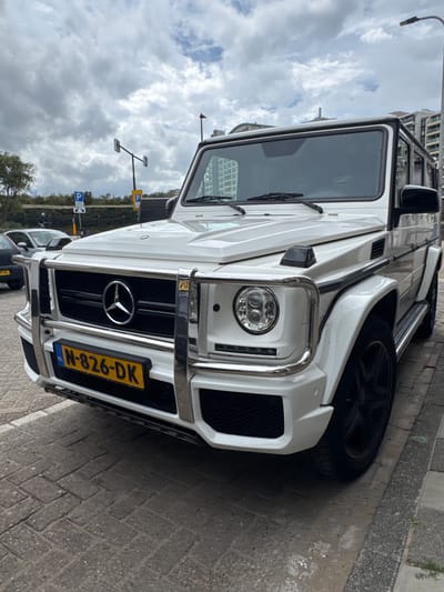 An outdoor, eye-level shot captures a white Mercedes-Benz G-Wagen (G-Class) SUV parked on what appears to be a paved street or sidewalk. The image is oriented vertically, showing the vehicle from the front-right quarter, with its front end angled towards the viewer.
The G-Wagen features a prominent chrome bull bar around the grille and headlights, round LED headlights, and the distinctive Mercedes-Benz star emblem centered on its black grille. A Dutch license plate, "N-826-DK," with the "NL" country identifier, is visible on the front bumper. The vehicle is white with black trim details, including its multi-spoke wheels.
Behind the G-Wagen, another darker vehicle is partially visible. The background features a partly cloudy sky, with several buildings, streetlights, and parking signs visible. One parking sign is yellow with a "P" symbol, and another blue sign also shows a "P." Green foliage, possibly bushes or trees, lines part of the background. The ground is paved with brick-like patterns on the sidewalk and a darker asphalt-like surface for the road.
The scene depicts a daytime setting with an overcast sky in Noordwijk, Netherlands.