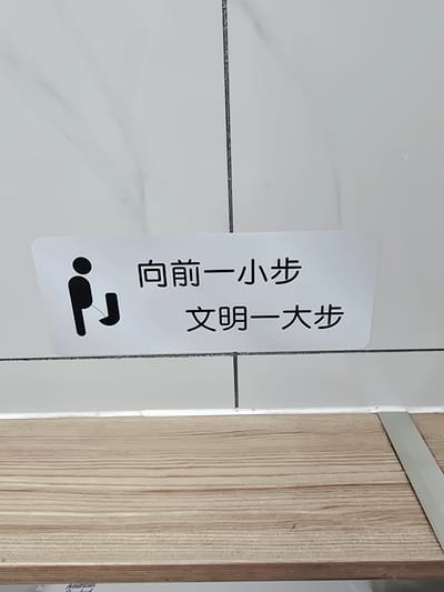 A sign with a pictogram of a man urinating and Chinese text is affixed to a tiled wall. The text reads "向前一小步 文明一大步", which translates to "One small step forward, a great step for civilization". The sign is white with black text and graphics. Below the sign is a wooden shelf. The environment appears to be a restroom.