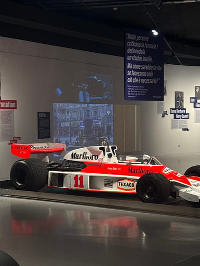 The image displays a vintage Formula 1 race car, predominantly white and red, featuring the number "11" on its side, set within an indoor exhibition space, possibly a museum in Turin, Italy.
**Subjects and Objects:** The central subject is the F1 car, detailed with sponsor logos such as "Marlboro" (prominently on the nose and sidepods), "TEXACO," "CHAMPION," "Lockheed," and "GOODYEAR." The name "James Hunt" is visible near the cockpit. Surrounding the car are various informational displays. A large, dark banner hangs, featuring a quote by Niki Lauda in both Italian and English. Other smaller, wall-mounted signs name individuals like "Ian Fleming" and "Dame Barbara Mary Quant," alongside another sign with "Coronation" and "TOO FAST TO LIVE TOO YOUNG TO DIE." A large, light-colored wall in the background shows a projected image, possibly an urban scene.
**Scene and Setting:** The setting is an indoor exhibition or museum. The car rests on a light-colored floor, possibly polished concrete, with reflections visible. The space is artificially lit, with spotlights illuminating the displays. The overall ambiance is clean and modern, typical of a professional exhibit.
**Activity or Event:** The image captures a static display, suggesting a historical or cultural exhibition dedicated to motorsport, specifically Formula 1, and possibly broader figures of influence. Visitors would engage by viewing the race car and reading the accompanying historical and biographical information.
**Notable Details:** The image is rotated 90 degrees clockwise. The Niki Lauda quote reads: "Molte persone criticano la Formula 1 definendola un rischio inutile. Ma come sarebbe la vita se facessimo solo ciò che è necessario?" and "A lot of people criticize Formula 1 as an unnecessary risk. But what would life be like if we only did what is necessary?". The juxtaposition of the F1 car with quotes and names of diverse public figures suggests a thematic exhibition exploring risk, ambition, or cultural impact. The time of day is not discernible due to the indoor setting and artificial lighting.