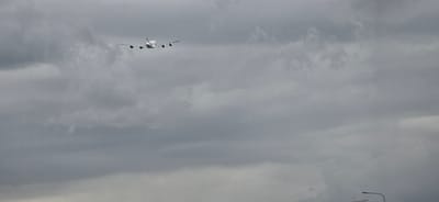 An airplane, identified as an Airbus A380 based on its distinctive four engines and double-deck configuration, is captured in mid-flight against a heavily overcast, grey sky. The aircraft is positioned in the upper left quadrant of the frame, viewed from the rear as it ascends or travels away from the viewer. The clouds are dense and uniform, with no visible breaks or patches of blue sky. The lighting conditions suggest a diffused light source, characteristic of an overcast day. In the lower right corner of the image, two streetlights with curved arms are partially visible, their presence indicating an urban or suburban environment. This scene is likely captured in the vicinity of Amsterdam Airport Schiphol (AMS), as the A380 is a wide-body airliner that operates on select routes and Schiphol is a major international airport in the Netherlands. Given the context of Hoofddorp being a town adjacent to Schiphol, this image was likely taken from Hoofddorp, Netherlands.