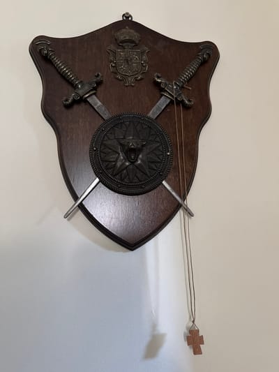 A wooden shield is mounted on a light-colored wall. Two decorative metal swords are crossed on the face of the shield, with their hilts pointing towards the top corners and blades angled downwards. Centered between the swords is a circular metal emblem, also decorative, depicting a stylized star with a face in the center. Above the swords and emblem, a smaller crest with a crown is affixed to the shield. A thin string hangs from the right sword, extending downwards and ending in a small wooden cross pendant. The shadow of the shield and its objects is cast on the wall to the lower left. The scene is devoid of people and activities, presenting a static display. This image was taken in Vnukovo, Russia.