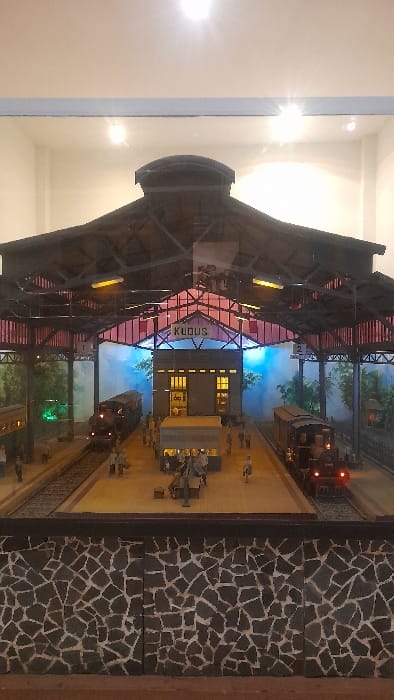 The image displays a detailed miniature scale model of a train station diorama, observed through a clear glass enclosure, located in a building in Glantengan, Indonesia. The central focus is a multi-track railway station with a prominent sign reading "KUDUS" positioned beneath a large, dark, arched canopy supported by an intricate red-painted metal framework.
The diorama includes a two-story station building featuring illuminated yellow windows and closed entrance doors. Two parallel railway tracks run through the station. On the left track, a dark-colored train with illuminated windows and a green front light is positioned. On the right track, a dark-colored steam locomotive with a single red headlight is visible.
A wide central platform separates the two tracks and is populated by numerous miniature human figures. These figures are depicted standing, walking, or seated on benches, some with miniature luggage nearby. One figure appears to be pushing a cart. Smaller platforms flank both tracks. The background consists of painted scenery depicting green trees and foliage, with a section of bright blue illumination on the right.
The immediate foreground of the image reveals the top edge of a display case's base or stand, constructed from an irregular mosaic pattern of dark and light grey stone pieces. Reflections of two overhead light sources are visible on the clear glass pane.