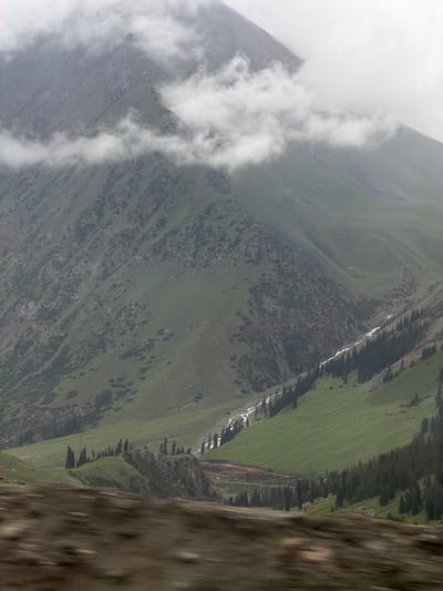 The image depicts a majestic mountain landscape in Kyrgyzstan. In the foreground, a blurred area suggests motion, possibly from a vehicle. The mid-ground features a vibrant green valley with a winding river or stream flowing through it, bordered by a dense line of tall, dark green coniferous trees. The terrain then ascends into rugged, green mountainsides dotted with rocky outcrops and patches of vegetation. Higher still, dramatic clouds partially obscure the peaks of the mountains, creating an ethereal and atmospheric effect. The sky is overcast, contributing to the subdued lighting of the scene. The overall impression is one of natural grandeur and serene wilderness.