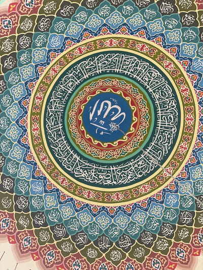 The image showcases intricate Islamic art, featuring a central circular design. Arabic calligraphy in white adorns concentric rings, with the name of Allah prominently displayed in the center on a blue background. The surrounding areas are filled with decorative geometric patterns and repeating calligraphic elements in various shades of blue, green, and brown. A silver chain is visible, likely attached to the central piece. The design reflects the symmetry and detail characteristic of Islamic art.