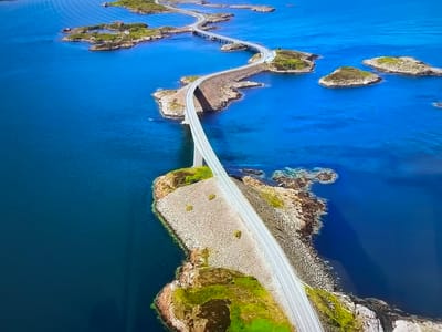 The image depicts the Atlantic Road in Norway. The road is a winding sequence of bridges and causeways connecting islands and skerries. The water is a deep blue, and the islands are rocky with patches of green vegetation. There is no evidence of the location being in Basingstoke and Deane, United Kingdom.