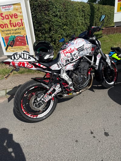 A motorcycle, possibly a Yamaha MT-07, is parked on asphalt in Much Dewchurch, United Kingdom. The motorcycle is white with red and black decals and graphics, including the words "Taster" and "Than". It has a black helmet and white riding gloves resting on the seat and a yellow and blue helmet placed on the ground behind it. A promotional sign for "Shell GO+ Rewards" is visible to the left of the motorcycle. The background consists of a green hedge and a brick wall. A shadow of a person is cast on the asphalt in the foreground.