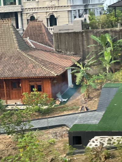 The image depicts an outdoor scene in Babakan Madang, Indonesia, featuring traditional Javanese-style wooden houses with clay tile roofs. In the foreground, a modern structure with a green roof and a black facade is visible, with a white decorative wall element. A paved pathway leads through the scene, bordered by dirt and vegetation. Banana trees and other green foliage are present. In the middle ground, smoke is emanating from a small fire near the wooden houses. There are no people clearly visible in the image. The background shows a large, multi-story building with ornate architectural details, likely part of a larger complex. The overall setting appears to be a hillside with a mix of traditional and modern architectural styles.