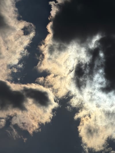 The image is a close-up, low-angle shot of dramatic, dark clouds illuminated by sunlight from behind. The clouds are a mix of deep grays and whites, with some areas appearing almost black while others are brightly lit and translucent. The sunlight creates a glowing effect, highlighting the edges and undersides of the clouds and casting luminous trails through the darker masses. There are no people, objects, or specific city landmarks visible. The scene is purely focused on the atmospheric conditions of the sky. This image was taken in Chirchik, Uzbekistan.