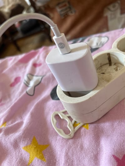 A white USB charging adapter, with a grey infinity symbol logo, is plugged into a dirty, white electrical socket. The socket is positioned on a pink fabric surface, which is adorned with yellow stars and white cloud-like patterns. A white USB cable extends from the adapter, curving upwards and out of the frame. The background is blurred, showing a patterned textile in shades of brown and white. The scene appears to be indoors, possibly on a bed or sofa. The location is not specified in the image or context.