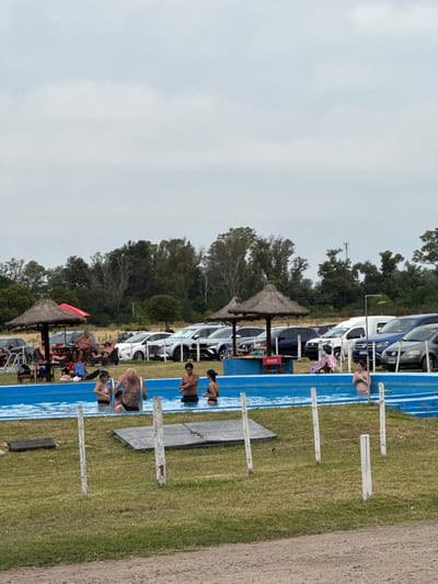 A waterside scene shows people enjoying a long, narrow, blue pool. Some people are in the water, while others lounge on the grass alongside it. Several thatched umbrellas offer shade, and a row of parked cars lines the edge of the area. The sky is overcast, suggesting a cloudy day. The scene exudes a relaxed, recreational atmosphere.