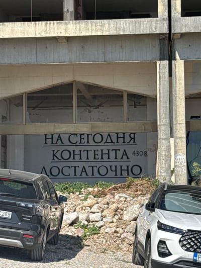 The image shows an unfinished concrete building in Perazića Do, Montenegro. Large, dark lettering is painted on the building's facade, reading "НА СЕГОДНЯ КОНТЕНТА ДОСТАТОЧНО" which translates to "ENOUGH CONTENT FOR TODAY." The building appears to be under construction or renovation, with exposed concrete structures and large window openings.
In the foreground, two cars are parked on a gravel surface. The car on the left is a black Suzuki S-Cross, and the car on the right is a white SUV. The ground is covered with gravel and scattered rocks. Vegetation, including small bushes and dry weeds, is visible around the cars and the building. Graffiti is present on one of the concrete columns to the right of the text, depicting a cartoonish figure and some unclear symbols. The overall scene suggests a public space, possibly a parking area or the entrance to a commercial or residential building, in a state of disrepair or development.