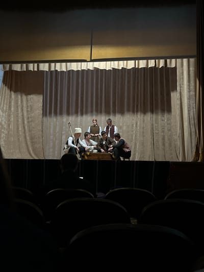 The image shows a group of approximately 7-10 people, possibly actors, gathered on a stage in Voiniagovo, Bulgaria. They are in costume, suggesting a play or performance. The stage has a draped curtain backdrop. Seating is visible next to the stage. The setting appears to be inside a theater. One person is holding a staff.