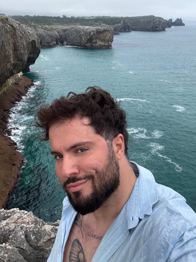 A man with dark, curly hair and a beard takes a selfie on a cliff overlooking the ocean in Llanes, Spain. He is wearing a light blue, pinstriped button-down shirt. A tattoo is visible on his right forearm, with the word "LIBERTAD" (freedom in Spanish) clearly discernible. The sea is a deep teal color, with white waves crashing against the rocky shore below. The cliffs are rugged and covered in greenery. The sky is overcast, suggesting a cool or possibly rainy day. The man's expression is relaxed and he is looking directly at the camera, smiling slightly. The orientation of the photo suggests it was taken at an angle.