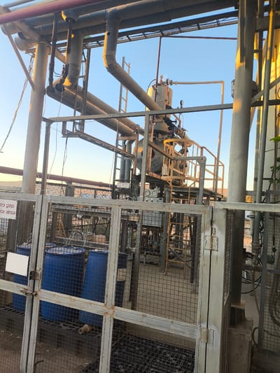 This media file depicts an outdoor industrial scene, likely in Dimona, Israel, during daylight hours, possibly late afternoon given the warm lighting. The setting is an industrial facility with a complex network of pipes, tanks, and metal structures.
Prominently featured are large blue barrels, some partially obscured by a metal fence. These barrels appear to be containers for liquids, possibly related to the industrial processes occurring on-site. The fence itself is made of metal mesh and frames, showing signs of weathering and rust.
Above the barrels, the scene reveals more industrial equipment. A tall, central structure houses a vessel with various gauges and pipes, suggesting a processing or storage unit. Numerous pipes of varying sizes and materials, some painted red, snake across the frame, supported by metal scaffolding. A ladder can be seen attached to one of the structures, indicating access points for maintenance.
The sky is clear and blue, indicating fair weather. The overall impression is one of a functional, working industrial site, with a focus on the infrastructure and equipment rather than any human activity.
There is some visible text in Hebrew on a white label attached to the fence. While not entirely clear, it appears to be a warning or informational sign.