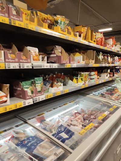The image depicts a well-lit supermarket aisle. The camera is positioned to capture shelves filled with various food products, including packaged goods and items in refrigerated display cases. On the shelves above, there are boxes of snacks, sauces, and other grocery items. Prominently displayed are yellow price tags. In the foreground, a refrigerated display case is filled with frozen food items, including meat products, packaged in plastic. The overall environment suggests a commercial setting for food retail. The location is stated to be Ходаево, Russia.
