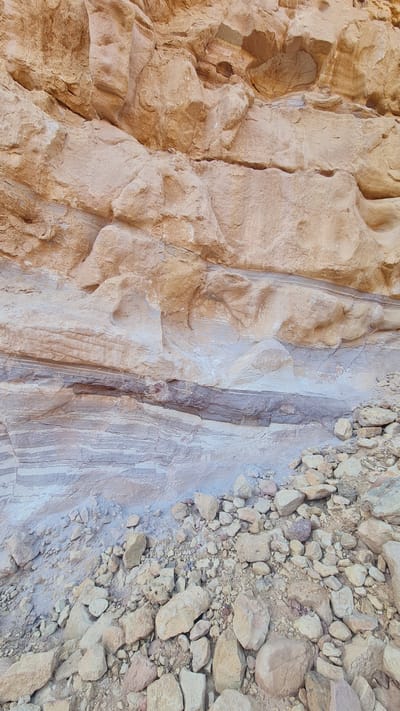 The image depicts a close-up view of a rock formation in Hevel Eilot Regional Council, Israel. The formation is composed of three distinct sections. The leftmost section is a rough, textured rock face with a tan color. The central section is smoother and lighter in color, with visible vertical striations. The rightmost section consists of loose rocks and pebbles, also in a tan color. The composition is a ground-level shot, focusing on the texture and variation in the rock. There are no people visible.