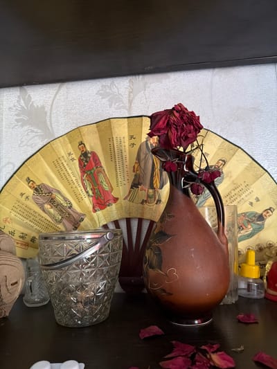 A still life scene is depicted on a dark shelf. In the foreground, a clear, faceted glass ice bucket with a metal handle is prominent. To its left, partially visible, is a light-colored, possibly ceramic, decorative object. Behind the ice bucket, a large, yellow, folding fan is displayed, decorated with images of figures in traditional attire and Chinese script. A reddish-brown ceramic vase, adorned with painted floral motifs, stands to the right of the fan. A single, dried, dark red rose is placed in the vase. Several dried rose petals are scattered on the shelf in front of the vase and ice bucket. The background is a textured, light-colored wallpaper with a floral pattern. The scene is lit from the front, casting soft shadows. The overall impression is of a curated display of decorative items. The location of this scene is Shepsi, Russia.