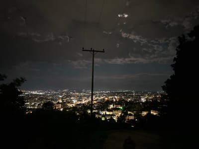 The image depicts a night scene in Xanthi, Greece, overlooking the city from an elevated position. The foreground is dark with silhouetted trees on either side, framing the view. A wooden utility pole stands prominently in the center of the frame, with power lines extending upwards towards the sky. The city of Xanthi stretches out in the midground, a dense collection of buildings illuminated by streetlights and windows. Beyond the city, the horizon is visible under a cloudy sky, where a glimpse of the moon can be seen. A faint shadow of a person appears to be visible at the bottom center of the frame.