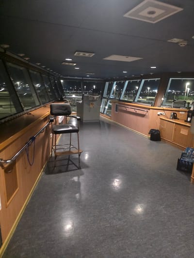 The image depicts the interior of a vessel's bridge or control room at night. The room is spacious with a dark gray ceiling containing recessed lighting and square decorative panels. Large windows line the perimeter, offering a view of a brightly lit cityscape or port, possibly Brindisi, Italy, with numerous streetlights and building lights visible.
In the foreground, a dark gray, textured floor reflects some of the overhead lighting. To the left, a wooden paneling lines the wall, with a chrome handrail attached. A blue rope is looped through the handrail. A black leather captain's chair with a metal frame and wooden footrest stands near the wall.
Further into the room, a lighter-colored wooden cabinet unit is visible along the right wall. On top of this unit, there is a black trash can, an electric kettle, and what appears to be a coffee maker. Next to the cabinet, a crate filled with bottled water is placed on the floor. The overall impression is of a functional, dimly lit operational space within a vessel.