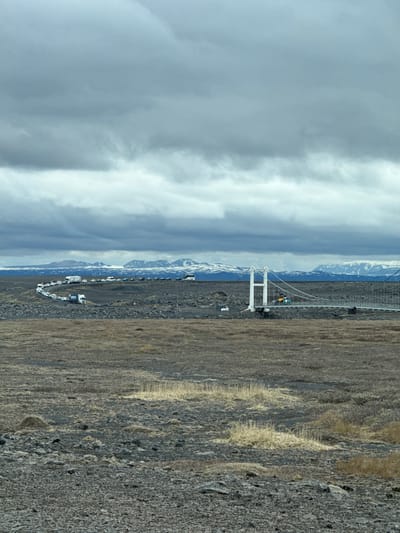 A panoramic shot captures a vast, rugged, and barren landscape under a heavily overcast sky, with a distinctive white suspension bridge serving as a central feature. The image, oriented horizontally, depicts a wide expanse of dark, rocky ground with sparse, dry, light brown vegetation in the foreground and stretching towards the middle ground.

In the mid-distance, a modern, white suspension bridge stands out against the dark terrain. It features two prominent A-frame towers supporting suspension cables, spanning what appears to be a fissure or a small canyon. Near the bridge, a small cluster of people, one wearing yellow, can be seen, suggesting pedestrian activity. To the left of the bridge, a long queue or parking line of various vehicles, including cars, vans, and possibly campervans, stretches into the distance. One blue and white vehicle, possibly a rescue or service truck, clearly displays the text "ICE-SAR" on its side, identifying it as belonging to the Icelandic Search and Rescue organization. This detail, along with the volcanic-looking terrain and distant mountains, strongly supports the given location context of Iceland.

In the background, a range of snow-capped mountains forms a dramatic horizon line, partially obscured by the expansive, thick grey clouds that dominate the upper half of the frame. The weather appears to be overcast daytime, with soft, diffused light, and the overall mood conveyed is one of a remote and expansive natural environment. The presence of numerous vehicles indicates that this bridge is likely a significant landmark or point of interest.