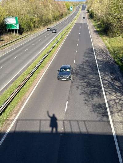 An elevated view shows a multilane highway with sparse traffic near Dacorum, United Kingdom. A dark car travels in the right-hand lane, casting a shadow resembling a person. Other vehicles are visible further down the highway. A sign indicates the direction and distance (1.3m) to Northchurchsted, Berkham, A4251, Wiggington, and a location named "Idington". Trees line the sides of the highway, casting shadows on the road.