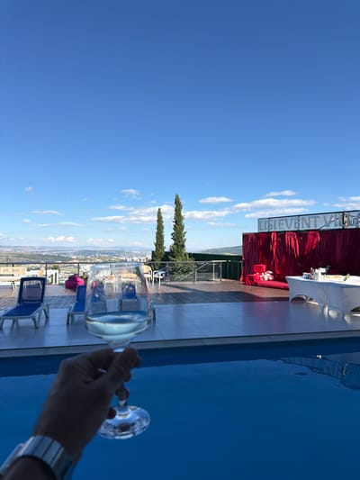 A person's hand, wearing a silver watch, holds a wine glass filled with white wine over a bright blue swimming pool. In the background, a wooden deck area overlooks a city with a distant mountainous horizon under a clear blue sky with scattered white clouds. The deck features several blue lounge chairs, a pink beanbag chair, and a stage area draped with red velvet curtains, adorned with floral arrangements and a white-topped table. A sign above the stage reads "LISIEVENT VILLA." The scene is set in Lisi, Georgia.