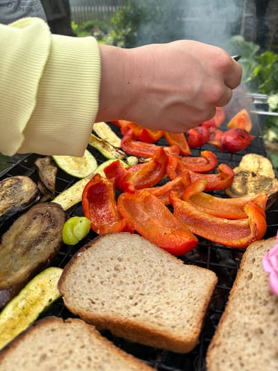 The image shows a close-up of a grill with various food items being cooked, including sliced red bell peppers, zucchini, eggplant, tomatoes, and slices of bread. A person's hand, wearing a light yellow long-sleeved shirt, is visible in the foreground, holding a skewer. Smoke is rising from the grill, suggesting it is actively in use. The scene appears to be outdoors, with greenery visible in the blurred background, hinting at a natural setting. The lighting suggests it is daytime, possibly late afternoon or early evening, given the warm tones of the food and the soft light. The overall impression is one of preparing a meal, likely a barbecue or outdoor gathering. No visible text is present in the image. The location context provided is Бронница, Russia.