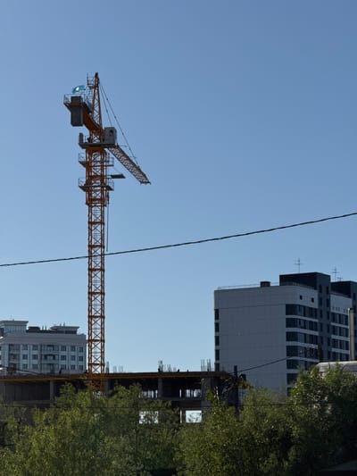The image depicts an outdoor scene with a clear blue sky. The main subjects are a tall orange construction crane and several modern buildings under construction. The crane is prominently positioned in the mid-ground, extending horizontally across the frame. One of its arms has a small blue and white flag attached. In the background, to the right of the crane, are the upper floors of a tall, grey and white building with many windows. To the far right, a dense green tree partially obscures another unfinished building. The overall setting suggests an urban environment undergoing development. The lighting and clear sky indicate a daytime setting, likely a sunny day. There is no visible text in the image. The image is oriented vertically, which may be an intentional artistic choice or a result of how the photo was captured. The scene evokes a sense of ongoing construction and urban growth.