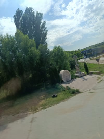 The image shows a roadside scene with a tall tree in the foreground, a cluster of green trees to the left, and a wide blue sky with white clouds. To the right of the trees, there is a paved area with a stone cross monument, a large round stone object, a small evergreen tree, and a concrete bench. Further in the background, there is a building with a blue awning, and beyond that, a hilly landscape with power lines. The ground in the foreground is a light-colored paved surface, with a patch of green grass to the left where a black tire is visible. The scene appears to be in Străisteni, Moldova, given the provided context.