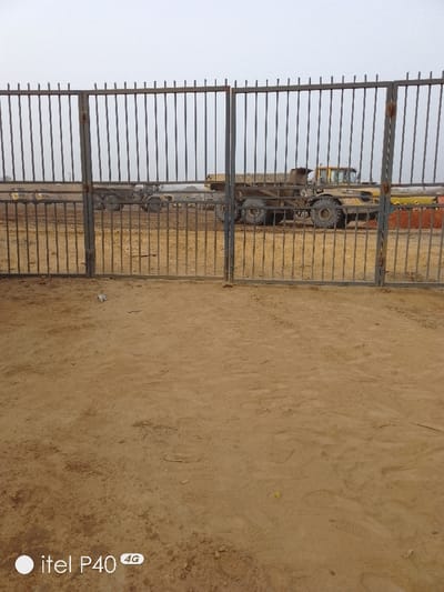 A fenced area of sandy ground is seen from behind a gate, revealing a construction site. Large yellow machinery, possibly dump trucks, are visible through the bars of the gate. The sky is overcast, suggesting a dull day. The ground in front of the gate is bare and sandy, likely the same material found within the fenced area. A logo from a phone is partially visible in the corner.