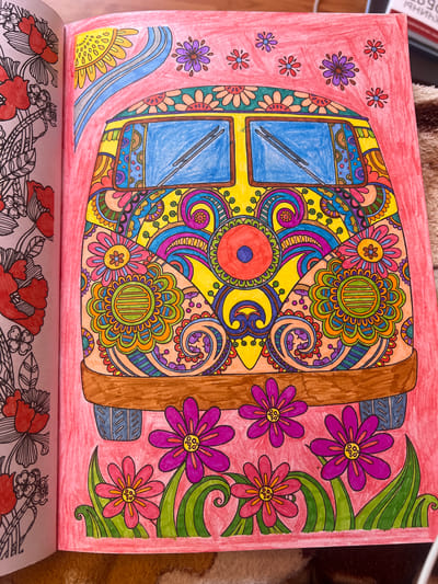 This is a close-up, overhead shot of an open coloring book page. The page features a vibrant, intricate mandala-style design with a variety of geometric shapes and floral motifs. The artwork is colored with bright, bold colors, including blue, yellow, orange, green, purple, and red.

The central element of the design appears to be a stylized representation of a window or a door, with two blue panes bordered by a brown frame. Surrounding this central area are numerous colorful flowers and swirling patterns. The background of the page is filled with a solid pink hue, and there are also some smaller, simpler flower designs scattered around the edges. To the right of the main design, there are several large purple and pink flowers with green leaves, also colored in.

Below this coloring page, the beginning of another page is visible, which contains a black and white line drawing of red flowers and decorative elements.

The overall impression is one of a finished coloring project, likely a page from an adult coloring book, given the complexity of the design. The lighting suggests it's daytime, and there are no obvious environmental clues about the weather or specific location beyond the implied indoor setting of a book. No text is clearly visible on the colored page itself.