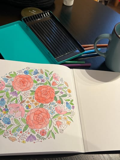 The scene depicts an overhead view of an indoor setting, likely a domestic environment in Tromsø, Norway, featuring materials for a coloring activity on a dark, reflective table surface. In the foreground, an open coloring book displays a partially completed mandala-style floral illustration on its left page. The design features three prominent red roses, surrounded by smaller flowers and foliage in shades of pink, blue, purple, yellow, and green. The right page of the book is blank white. In the midground, to the left, an open teal-colored metal pencil case with a black partitioned interior, currently empty of pencils, rests on the table. To the right of the pencil case, several colored pencils (brown, blue, green, purple) are scattered directly on the table. Further to the right, a light teal or grey-blue ceramic mug with a handle is visible. In the background, partially obscured by the pencil case, a dark green tin labeled "Epok No. 1 Ice Blue" and other indistinct objects are present. No individuals are visible.