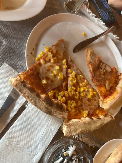 In Ribaritsa, Bulgaria, a white plate holds three slices of pizza with corn and melted cheese. A knife rests on the plate next to a single corn kernel. A glass ashtray filled with cigarette butts and ash sits to the lower right of the plate. A white napkin is placed beneath the plate and knife. A dark-colored phone is visible at the top right of the image. The scene is set on a wooden table covered with a woven placemat.