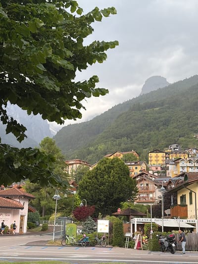 This media file shows a street scene in Molveno, Italy. The image is oriented vertically. In the foreground, lush green trees with large leaves dominate the left and bottom sections, framing the view.

Beyond the trees, a street runs horizontally across the middle and right side of the frame. On the right side of the street, several buildings are visible. One building features a sign that reads "GELATERIA - PASTI - CAFE DRINK." Another sign, partially obscured, says "Pizzico - BAR PIZZERIA GELATERIA."

Several objects are present on the street and sidewalk. A black motorcycle is parked near the building with the "GELATERIA" sign. Two bicycles are also visible further down the street, one leaning against a green structure that appears to be a bus stop or shelter.

Several people are present. A man in a dark shirt and jeans is leaning against a railing near the "GELATERIA" sign. Two people, one in a dark top and one in a dark shirt, are sitting on a bench outside the "Pizzico" establishment. Another person is walking in front of the "Pizzico" building.

In the background, a dense, green forest covers a steep mountainside, receding into the distance. The sky above is overcast and appears cloudy, suggesting either early morning or a cloudy day. The overall atmosphere is calm and suggests a leisurely day in a European mountain town.