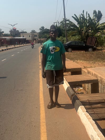The image depicts a daytime scene on a street in Aguata, Nigeria. A dark-skinned male subject stands centrally in the foreground, facing the camera directly. He wears a black beanie, a green short-sleeved t-shirt with a "NIKE" logo, dark shorts with white side stripes and a white drawstring, and dark brown flip-flops. His hands are at his sides.

To the subject's left is an asphalt road with a solid yellow line delineating the shoulder and white dashed lines marking lanes further left. A person on a motorcycle is visible in the mid-ground, traveling away from the camera. In the background along the road, several buildings of varying sizes and designs are present, alongside modern solar streetlights.

To the subject's right, an unpaved dirt shoulder borders the road, followed by a concrete drainage ditch. The ditch is partially covered by concrete slabs and wooden planks, and contains visible refuse, including plastic items. Beyond the ditch and shoulder, a black sedan (consistent with a 1990s-2000s model Toyota Camry) is parked on a dirt patch, facing left. The vehicle's trunk appears open or damaged, with several metal frames, possibly window grilles, resting against it. Lush green foliage, including banana trees, densely covers the area behind the car and extends into the background. A wooden utility pole with overhead wires is situated near the sedan. The sky is clear, and direct sunlight illuminates the scene, casting shadows.