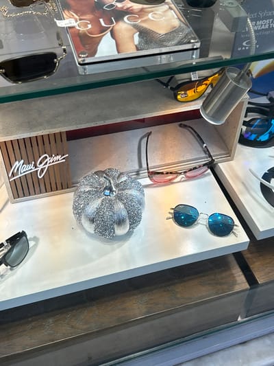 This media file captures an indoor retail display of sunglasses and accessories in Koridallos, Greece. The scene is presented from a slightly elevated, tilted perspective, looking down into a well-lit display case.
The primary subjects are several pairs of sunglasses. In the upper left, a pair with yellow/orange mirrored lenses is held by a metal stand with an integrated spotlight. To its right, a pair with reddish-pink frames lies flat. Below these, two pairs of roundish sunglasses with blue mirrored lenses are arranged vertically. Towards the bottom right, another pair of dark-lensed sunglasses is visible. Another dark pair is partially visible in the bottom left, below a promotional poster.
A notable decorative item is a silver, sparkly pumpkin, positioned centrally in the right half of the image, suggesting a festive or seasonal accent. Below it, a wooden slatted sign displays the "Maui Jim" logo in white script.
The display includes various surfaces: a light-colored shelf or counter, a grey-toned display box, and a rich brown wooden element on the right. A glass partition of a display case runs vertically on the left side.
Visible text includes "Maui Jim" on the wooden sign and "Gucci" on a poster featuring a woman's image, partially seen through the glass in the upper left. Other partial texts include "Meet Sphaer," "R MOST WEAR TO," "So wou'll forger y," and fragments suggesting "Oakley" (DAK Y).
There is no direct human activity, but the organized arrangement clearly indicates a retail setup for showcasing eyewear products. The lighting is bright, creating highlights on the sunglass lenses and the sparkly pumpkin.