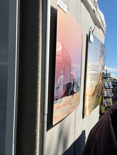The image shows an outdoor art display, likely at a street fair or market, in the Village of Sag Harbor, United States of America. Two large, curved photographs are prominently displayed on a textured, light-colored wall. The photograph on the left features a warm-toned scene with a person in a white shirt standing in front of what appears to be a building or structure, with a pinkish hue cast over the image. The photograph on the right depicts a sunlit landscape, possibly a beach or water scene, with golden light reflecting on the surface. The photos are hung with thin wires, and one of them has a clothespin holding a small tag. In the background, to the right, a rack filled with colorful items, possibly merchandise, is visible. A person's shoulder and upper arm, wearing a dark garment, are in the foreground on the right side of the image, casting a shadow on the wall. The scene is illuminated by natural sunlight, creating strong contrasts and shadows. The sky above is a clear, bright blue.