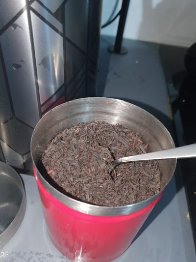 A close-up, eye-level shot shows a red metal container filled with loose-leaf black tea. A silver spoon is partially submerged in the tea. To the left of the red container is a silver lid, also appearing to be made of metal, and further to the left, a portion of a tall, silver thermos with a geometric pattern is visible. The objects are arranged on a grey surface, which may be a countertop or table. The background shows a blurred dark vertical object and a lighter grey area, possibly a wall or another surface. The lighting suggests an indoor environment, with a bright light source casting reflections on the metal objects. There are no people or animals visible in the image. The scene is situated within Kheyrabad, Iran.