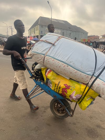 In Egan, Nigeria, an outdoor scene depicts a male individual pushing a heavily loaded, single-wheeled blue metal pushcart on a paved road. The man, with dark skin, wears a black t-shirt, light-colored shorts, and dark slide sandals, gripping the cart's handles while looking forward and slightly left. The cart carries a large, light-colored mattress or bundle secured with straps on top, a smaller dark patterned bundle underneath, and a large yellow sack labeled "LONG GRAIN PARBOILED RICE PEARL" at the bottom. Another male individual, partially visible behind the cart's load, walks alongside, looking right. In the background, a two-story building with a grey roof and light-colored walls stands, displaying a sign reading "MARO" and "NATIONAL BANK OF NIGERIA." A black street lamp pole extends upwards. The sky appears overcast. Several other indistinct figures are visible in the distant background, suggesting an active urban or market environment.