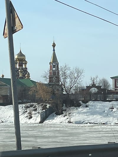 The image depicts a view in Норья, Russia, showcasing a Russian Orthodox church complex in winter. The architecture is characterized by tiered structures with gold domes and a bell tower. The ground is covered in snow, with icicles hanging from buildings and structures. Power lines are visible in the foreground, partially obscuring the view. Bare trees suggest a cold season. The overall scene is a mix of historical architecture and winter landscape.